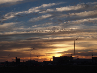 [The sun just below the horizon backlights the buildings and street light posts in the distance. Above the many scattered clouds is blue sky. The clouds are white at the top of the image, but shades of grey, white, and yellow closer to the horizon.]